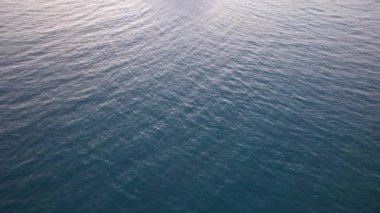 Günbatımında okyanusta yalnız bir ada duruyor. İnsansız hava aracının görüntüsü. Etrafındaki küçük dalgalar okyanus yüzeyinde dalgalanmalar yaratıyor. Su siyah bir ayna gibidir. Pembemsi kara bulutlar