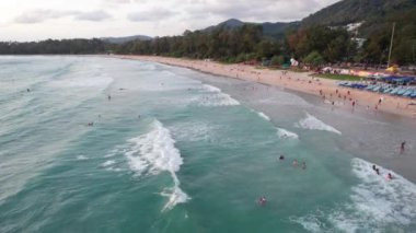 Gün batımında plajda sörf yapmak. İnsansız hava aracının görüntüsü. Küçük dalgalar köpük yaratır. Adam sahile giden bir tahtanın üzerinde uçuyor. Su, güneş ışınlarını yansıtır. İnsanlar banyo yapıyor. Phuket