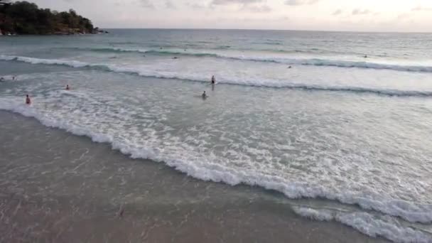 日没時にビーチの近くでパドルサーフィン ドローンからの眺め 小さな波が泡を作ります 男は岸にボード上を飛んでいる 水は太陽の光を反射する 人々は入浴している プーケット — ストック動画