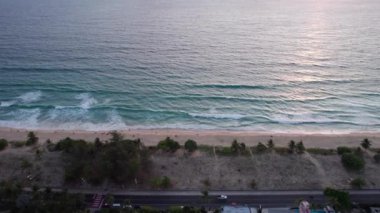 Deniz üzerinde bulutlu kırmızı bir günbatımı manzarası. Yeşil ada Phuket. Kumsalda sarı kum. İnsanlar dinleniyor. Kıyıya yakın dalgalar. Oteller var. Suyun üzerindeki turuncu güneş ışınları