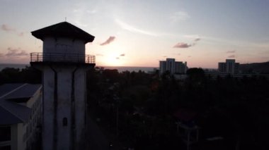 Gün batımında Phuket Adası manzaralı su kulesi. Turuncu güneş, deniz ve bulutlar. Kulenin etrafında yeşil palmiye ağaçları, oteller ve yüzme havuzları var. İnsanlar banyo yapıyor. Karon, ulaşımı geç.