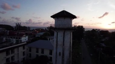 Gün batımında Phuket Adası manzaralı su kulesi. Turuncu güneş, deniz ve bulutlar. Kulenin etrafında yeşil palmiye ağaçları, oteller ve yüzme havuzları var. İnsanlar banyo yapıyor. Karon, ulaşımı geç.