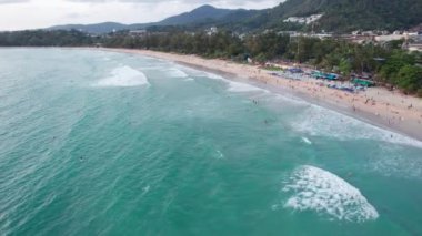 İnsanlar plajda dinleniyor. İnsansız hava aracının görüntüsü. Büyük dalgalar köpük yaratır. Kıyıda palmiye ağaçları yetişir, büyük taşlar yatar. Bazı yerlerde güneş pansiyonları ve kafeler var. Akşam okyanusu. Phuket