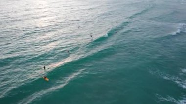 Gün batımında plajda sörf yapmak. İnsansız hava aracının görüntüsü. Küçük dalgalar köpük yaratır. Adam sahile giden bir tahtanın üzerinde uçuyor. Su, güneş ışınlarını yansıtır. İnsanlar banyo yapıyor. Phuket