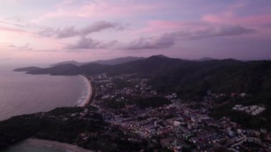 Yeşil tepeler ve denizin ortasındaki bir adada gün batımı. Gökyüzü ve bulutların pembe-mor rengi. Dalgalı devasa plajlar. Deniz manzarası ve akşam şehri Karon Phuket Adası, Tayland Işıkları açık.