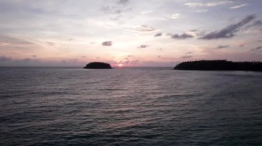 Günbatımında okyanusta yalnız bir ada duruyor. İnsansız hava aracının görüntüsü. Etrafındaki küçük dalgalar okyanus yüzeyinde dalgalanmalar yaratıyor. Su siyah bir ayna gibidir. Pembemsi kara bulutlar