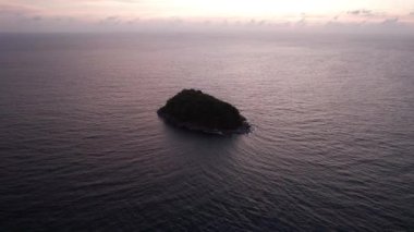 Günbatımında okyanusta yalnız bir ada duruyor. İnsansız hava aracının görüntüsü. Etrafındaki küçük dalgalar okyanus yüzeyinde dalgalanmalar yaratıyor. Su siyah bir ayna gibidir. Pembemsi kara bulutlar