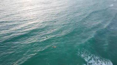 Gün batımında plajda sörf yapmak. İnsansız hava aracının görüntüsü. Küçük dalgalar köpük yaratır. Adam sahile giden bir tahtanın üzerinde uçuyor. Su, güneş ışınlarını yansıtır. İnsanlar banyo yapıyor. Phuket