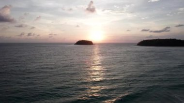 Günbatımında okyanusta yalnız bir ada duruyor. İnsansız hava aracının görüntüsü. Etrafındaki küçük dalgalar okyanus yüzeyinde dalgalanmalar yaratıyor. Su siyah bir ayna gibidir. Pembemsi kara bulutlar