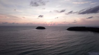 Günbatımında okyanusta yalnız bir ada duruyor. İnsansız hava aracının görüntüsü. Etrafındaki küçük dalgalar okyanus yüzeyinde dalgalanmalar yaratıyor. Su siyah bir ayna gibidir. Pembemsi kara bulutlar