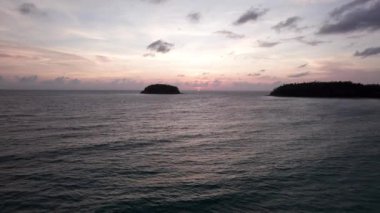 Günbatımında okyanusta yalnız bir ada duruyor. İnsansız hava aracının görüntüsü. Etrafındaki küçük dalgalar okyanus yüzeyinde dalgalanmalar yaratıyor. Su siyah bir ayna gibidir. Pembemsi kara bulutlar