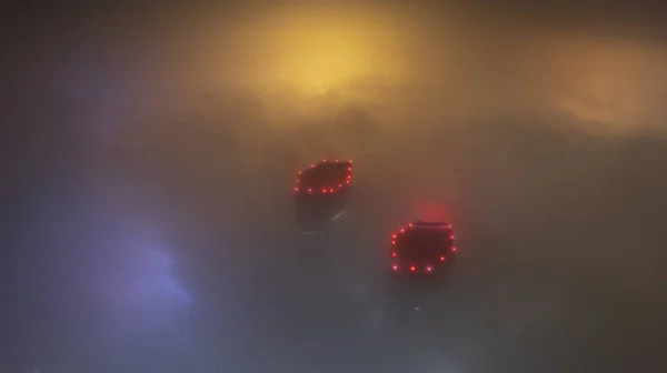 Buildings with bright lights in the night fog. Gemini is like a spaceship. Bright red lights on the roof, blue-yellow illumination on the fog. The view from the drone. Almaty. Lanterns are visible