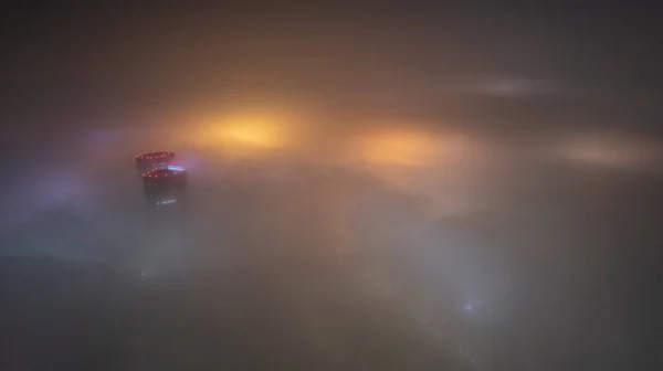 Buildings with bright lights in the night fog. Gemini is like a spaceship. Bright red lights on the roof, blue-yellow illumination on the fog. The view from the drone. Almaty. Lanterns are visible