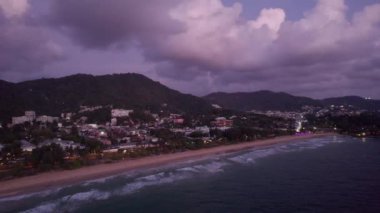 Phuket Adası 'nın insansız hava aracı görüntüsü. Karon Sahili. Dalgalı karanlık deniz suyu. Yüksek yeşil tepeler ve gri bulutlar. Şehrin ışıkları ve geçen arabalar parlıyor. Gün batımı. Tepedeki Büyük Buda