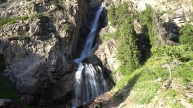 Yeşil bir ormanda kayaların arasında yüksek bir şelale. Nehir akıyor. Büyük taşlar var. Uzun kozalaklı ağaçlar ve yeşil otlar. Şelale şelalesi. Yosun büyür. Küçük bir dron uçuyor. Burhan Bulak