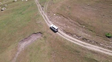 Beyaz bir SUV nehir boyunca uzanan bir vadi boyunca bir dağ yolunun üstesinden gelir. Uçurumlu yeşil alanlar. Dik uçurumlar ve yoldaki büyük kayalar. Keskin bir iniş ve tepe manzarası. Noel ağaçları