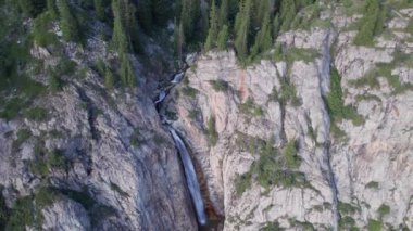 Bir dağ geçidinde yüksek bir şelale çağlıyor. Kozalaklı ağaçlar, Noel ağaçları ve otlar yetişir. Nehir akıyor. Yürüyüş yolları. İHA 'nın üst görüntüsü. Gün batımı. Burkhan Bulak Şelalesi, Kazakistan