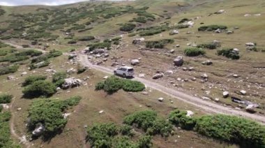 Beyaz bir SUV nehir boyunca uzanan bir vadi boyunca bir dağ yolunun üstesinden gelir. Uçurumlu yeşil alanlar. Dik uçurumlar ve yoldaki büyük kayalar. Keskin bir iniş ve tepe manzarası. Noel ağaçları