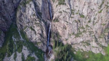 Bir dağ geçidinde yüksek bir şelale çağlıyor. Kozalaklı ağaçlar, Noel ağaçları ve otlar yetişir. Nehir akıyor. Yürüyüş yolları. İHA 'nın üst görüntüsü. Gün batımı. Burkhan Bulak Şelalesi, Kazakistan