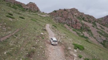 Beyaz bir SUV nehir boyunca uzanan bir vadi boyunca bir dağ yolunun üstesinden gelir. Uçurumlu yeşil alanlar. Dik uçurumlar ve yoldaki büyük kayalar. Keskin bir iniş ve tepe manzarası. Noel ağaçları