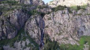 Bir dağ geçidinde yüksek bir şelale çağlıyor. Kozalaklı ağaçlar, Noel ağaçları ve otlar yetişir. Nehir akıyor. Yürüyüş yolları. İHA 'nın üst görüntüsü. Gün batımı. Burkhan Bulak Şelalesi, Kazakistan