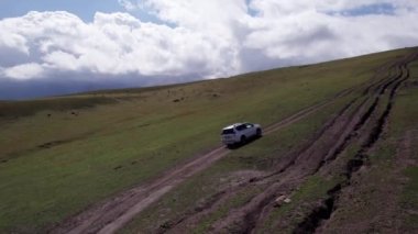 Beyaz bir SUV dağlarda tarlalardan geçiyor. Devasa beyaz bulutlardan, karlı dağ zirvelerinden ve yeşil tarlalardan oluşan bir dronun hava görüntüsü. Su birikintileri olan kirli bir off-road. Tekerleklerden damlıyor