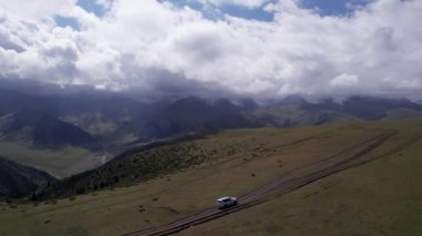Beyaz bir SUV dağlarda tarlalardan geçiyor. Devasa beyaz bulutlardan, karlı dağ zirvelerinden ve yeşil tarlalardan oluşan bir dronun hava görüntüsü. Su birikintileri olan kirli bir off-road. Tekerleklerden damlıyor