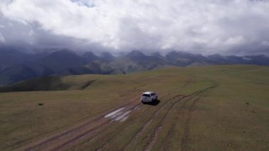 Beyaz bir SUV dağlarda tarlalardan geçiyor. Devasa beyaz bulutlardan, karlı dağ zirvelerinden ve yeşil tarlalardan oluşan bir dronun hava görüntüsü. Su birikintileri olan kirli bir off-road. Tekerleklerden damlıyor