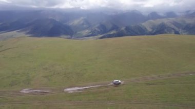 Beyaz bir SUV dağlarda tarlalardan geçiyor. Devasa beyaz bulutlardan, karlı dağ zirvelerinden ve yeşil tarlalardan oluşan bir dronun hava görüntüsü. Su birikintileri olan kirli bir off-road. Tekerleklerden damlıyor