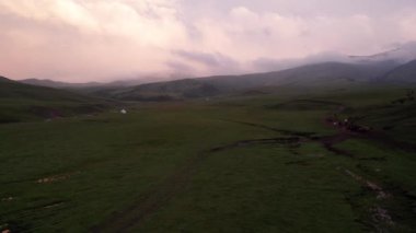 Yeşil tarlalı dağlarda pembe-mor gün batımı. Beyaz bir yurt var, bir dağ nehri akıyor. Kocaman bulutlar. Bir at sürüsü yürüyor. Çamur ve çadırın yanında bir SUV. Turistler toplanıyor