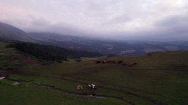 Yeşil tarlalı dağlarda pembe-mor gün batımı. Beyaz bir yurt var, bir dağ nehri akıyor. Kocaman bulutlar. Bir at sürüsü yürüyor. Çamur ve çadırın yanında bir SUV. Turistler toplanıyor