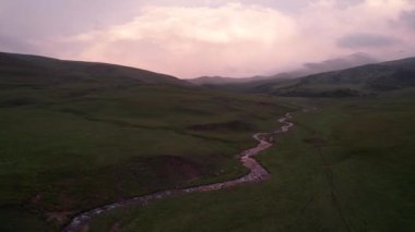 Yeşil tarlalı dağlarda pembe-mor gün batımı. Beyaz bir yurt var, bir dağ nehri akıyor. Kocaman bulutlar. Bir at sürüsü yürüyor. Çamur ve çadırın yanında bir SUV. Turistler toplanıyor