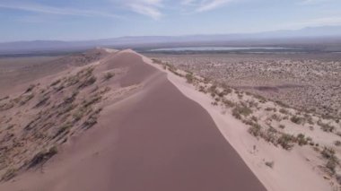 Bozkırın ortasında büyük bir kum tepeciği. Kocaman bir kum yığınının drone görüntüsü. Turistler yürüyor, manzaranın tadını çıkarıyorlar. Uzakta bir nehir akar ve çimenler büyür. Beyaz bulutlu gökyüzü