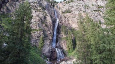 Bir dağ geçidinde yüksek bir şelale çağlıyor. Kozalaklı ağaçlar, Noel ağaçları ve otlar yetişir. Nehir akıyor. Yürüyüş yolları. İHA 'nın üst görüntüsü. Gün batımı. Burkhan Bulak Şelalesi, Kazakistan