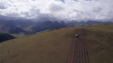 Beyaz bir SUV dağlarda tarlalardan geçiyor. Devasa beyaz bulutlardan, karlı dağ zirvelerinden ve yeşil tarlalardan oluşan bir dronun hava görüntüsü. Su birikintileri olan kirli bir off-road. Tekerleklerden damlıyor