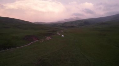Yeşil tarlalı dağlarda pembe-mor gün batımı. Beyaz bir yurt var, bir dağ nehri akıyor. Kocaman bulutlar. Bir at sürüsü yürüyor. Çamur ve çadırın yanında bir SUV. Turistler toplanıyor
