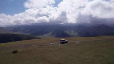 Beyaz bir SUV dağlarda tarlalardan geçiyor. Devasa beyaz bulutlardan, karlı dağ zirvelerinden ve yeşil tarlalardan oluşan bir dronun hava görüntüsü. Su birikintileri olan kirli bir off-road. Tekerleklerden damlıyor