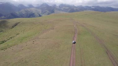 Beyaz bir SUV dağlarda tarlalardan geçiyor. Devasa beyaz bulutlardan, karlı dağ zirvelerinden ve yeşil tarlalardan oluşan bir dronun hava görüntüsü. Su birikintileri olan kirli bir off-road. Tekerleklerden damlıyor