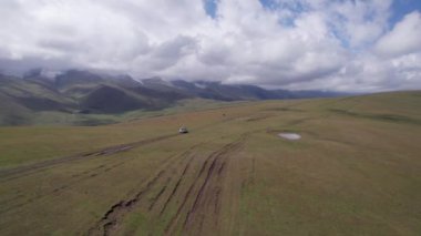 Beyaz bir SUV dağlarda tarlalardan geçiyor. Devasa beyaz bulutlardan, karlı dağ zirvelerinden ve yeşil tarlalardan oluşan bir dronun hava görüntüsü. Su birikintileri olan kirli bir off-road. Tekerleklerden damlıyor