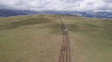 Beyaz bir SUV dağlarda tarlalardan geçiyor. Devasa beyaz bulutlardan, karlı dağ zirvelerinden ve yeşil tarlalardan oluşan bir dronun hava görüntüsü. Su birikintileri olan kirli bir off-road. Tekerleklerden damlıyor