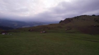 Yeşil tarlalı dağlarda pembe-mor gün batımı. Beyaz bir yurt var, bir dağ nehri akıyor. Kocaman bulutlar. Bir at sürüsü yürüyor. Çamur ve çadırın yanında bir SUV. Turistler toplanıyor