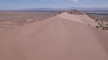 Bozkırın ortasında büyük bir kum tepeciği. Kocaman bir kum yığınının drone görüntüsü. Turistler yürüyor, manzaranın tadını çıkarıyorlar. Uzakta bir nehir akar ve çimenler büyür. Beyaz bulutlu gökyüzü