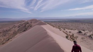 Bozkırın ortasında büyük bir kum tepeciği. Adam manzaranın tadını çıkarıyor. Kumda ayak izleri. Kocaman bir kum tepesi. Mavi gökyüzünde beyaz bulutlar. Çimenler bozkırda yetişir. Şarkı söyleyen Dune, Kazakistan