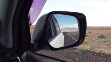 Arabanın içinden yan aynaya doğru bak. Araba bozkırın ortasında yol boyunca araba sürüyor. Kuru çalılar, direkler. Beyaz bir arabanın, bir yolun ve gökyüzünün yansımasında