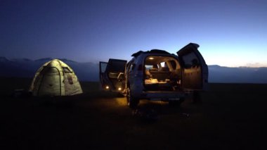 Gün batımında bir çadırın ve arabanın önünde kamp yapmak. Uzakta, güneş ve bulutlar ufukta kayboluyor, gökyüzündeki yıldızlar. Çadırda bir fener parlıyor, bacadan duman çıkıyor..
