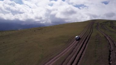 Beyaz bir SUV dağlarda tarlalardan geçiyor. Devasa beyaz bulutlardan, karlı dağ zirvelerinden ve yeşil tarlalardan oluşan bir dronun hava görüntüsü. Su birikintileri olan kirli bir off-road. Tekerleklerden damlıyor
