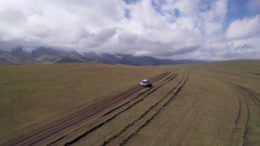 Beyaz bir SUV dağlarda tarlalardan geçiyor. Devasa beyaz bulutlardan, karlı dağ zirvelerinden ve yeşil tarlalardan oluşan bir dronun hava görüntüsü. Su birikintileri olan kirli bir off-road. Tekerleklerden damlıyor