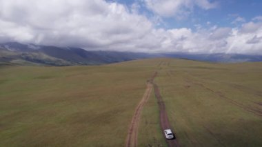 Beyaz bir SUV dağlarda tarlalardan geçiyor. Devasa beyaz bulutlardan, karlı dağ zirvelerinden ve yeşil tarlalardan oluşan bir dronun hava görüntüsü. Su birikintileri olan kirli bir off-road. Tekerleklerden damlıyor