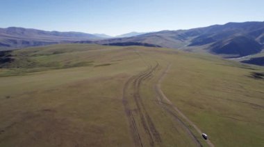 Beyaz bir SUV dağ yeşili tarlalardan geçiyor. Uzakta karlı zirveleri olan yüksek tepeler var. Kirli yol. Mavi gökyüzü. Bir köpek koşar ve çobanlık yapar. Dağlık arazi, bazen orman.