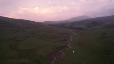 Yeşil tarlalı dağlarda pembe-mor gün batımı. Beyaz bir yurt var, bir dağ nehri akıyor. Kocaman bulutlar. Bir at sürüsü yürüyor. Çamur ve çadırın yanında bir SUV. Turistler toplanıyor